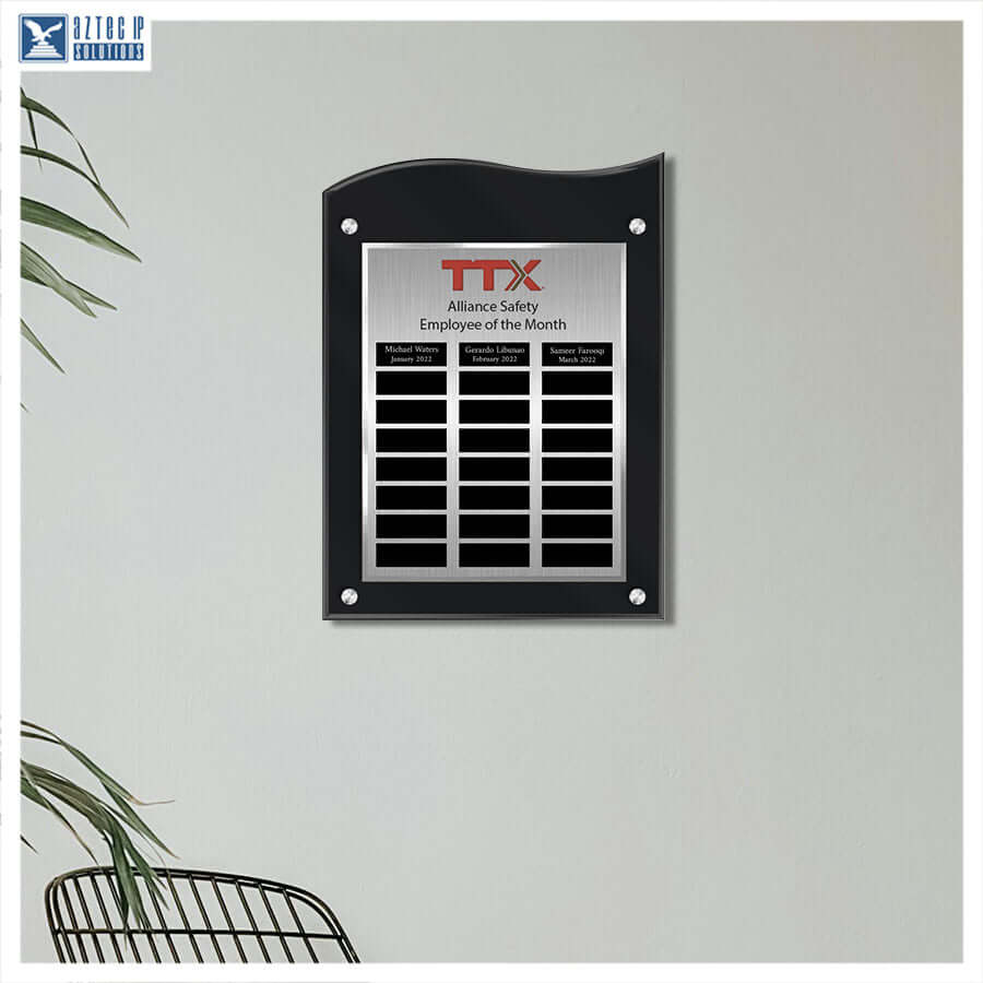 A black acrylic and stainless steel employee plaque mounted on a light gray wall, featuring the red and black TTX logo and three columns of name slots. A small TTX Solutions logo appears in the top-left corner, with a plant and chair partially visible at the edges.