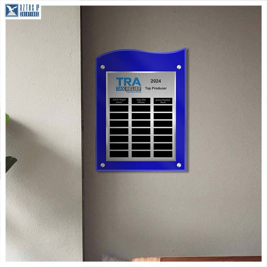A blue acrylic wall plaque with a silver center panel displaying 'TRA Taxpayers Trust 2024 Top Producer' and black name blocks, mounted on a beige wall.