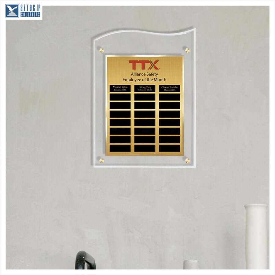 A gold and black acrylic employee of the month plaque mounted on a light gray textured wall. The plaque features the red and black TTX Alliance Safety logo at the top, followed by three columns of black nameplates, and is framed in a clear acrylic border with gold corner fasteners.