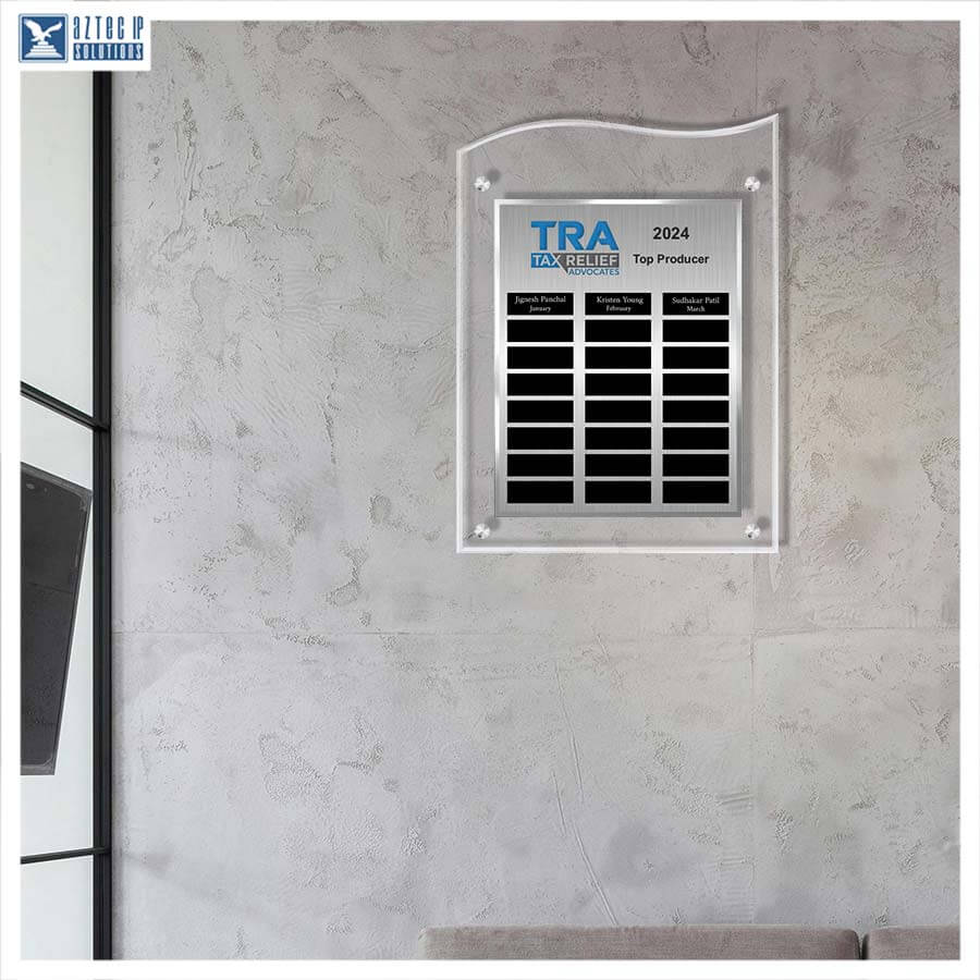 A clear acrylic wall plaque with a black grid of name slots and a blue 'TRA EXCELLENCE 2024 Top Producer' logo, mounted on a textured light gray wall.