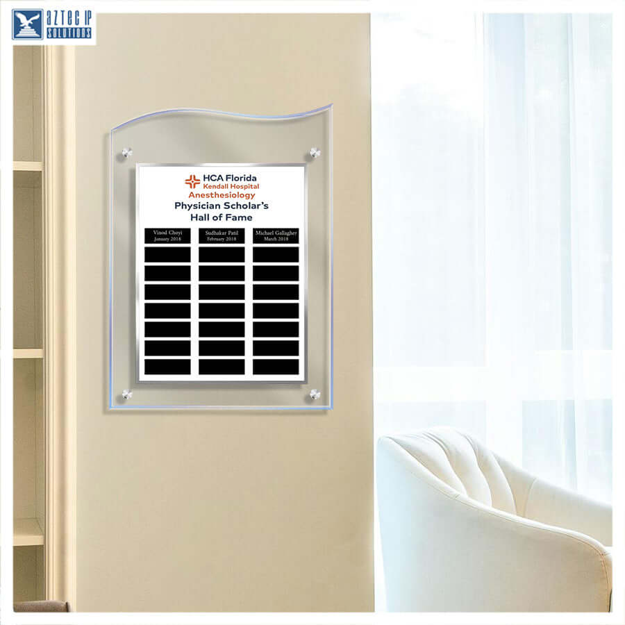A clear acrylic wall plaque with a printed white insert for HCA Florida Health System Physician Scholar's Hall of Fame, mounted on a beige wall in a bright interior setting with a chair and window visible.