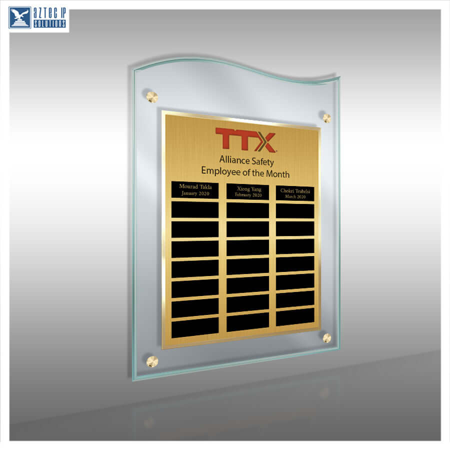 A wall-mounted acrylic and brushed gold plaque with a glass border, featuring the red TTX logo and black nameplates for monthly safety awards. The plaque is shown against a gray gradient background with a faint reflection below.
