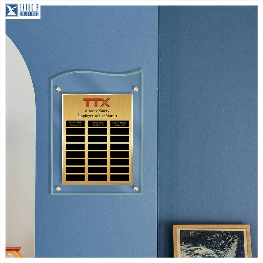 A gold and black TTX Alliance Safety Employee of the Month plaque mounted on a blue wall, with a small logo watermark in the top-left corner.